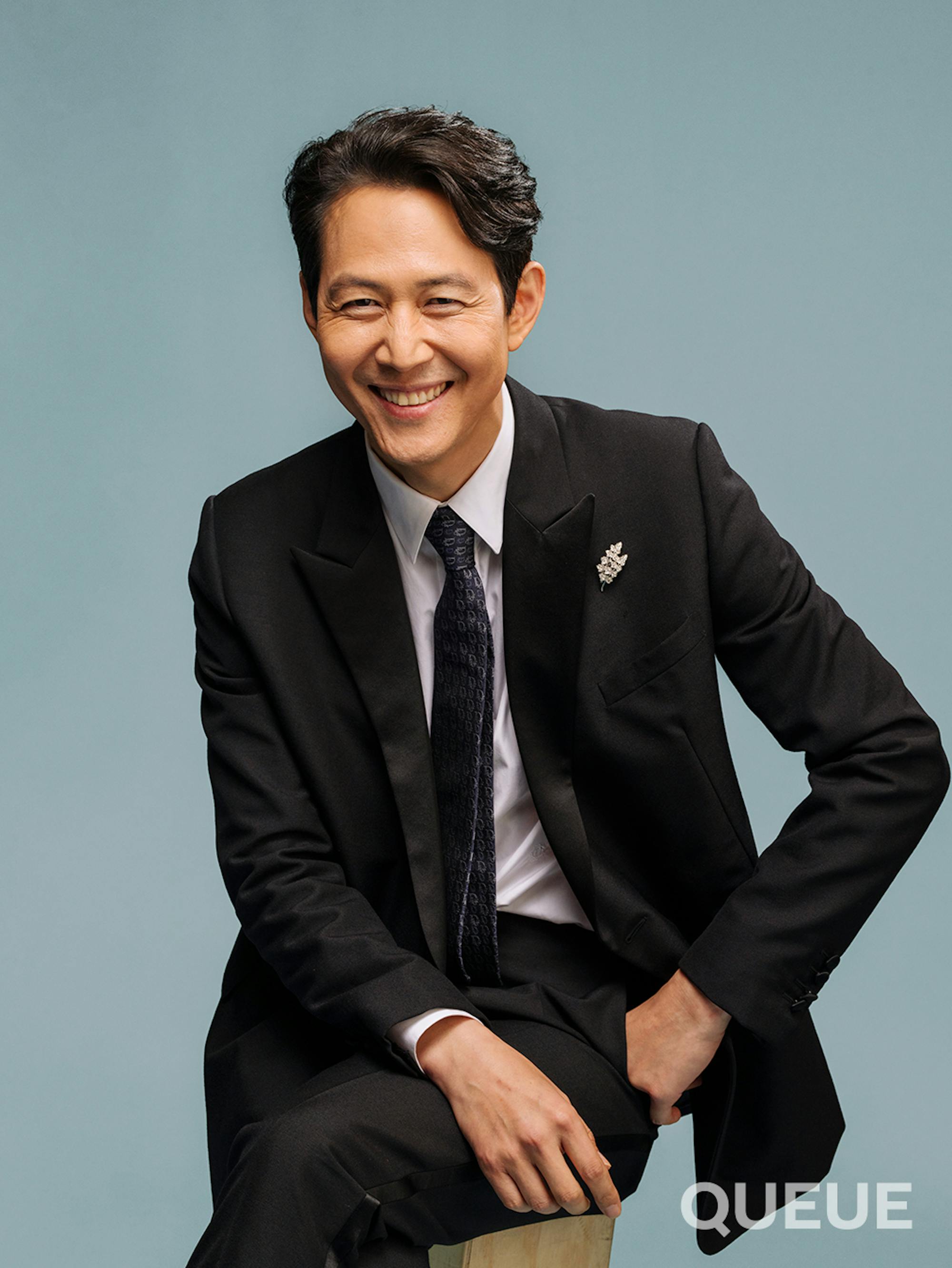 Lee Jung-jae in front of a gray-blue background smiling wide with one hand draped over his lap and the other at an angle going slightly into his pant pocket also wearing a sophisticated suit.