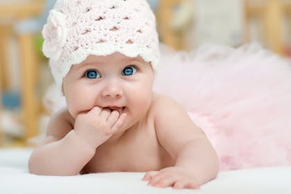 baby girl with blue eyes wearing hat lying on her stomach and sucks fingers