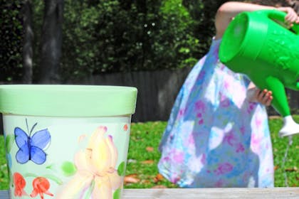 Girl pours water from a watering can next to a painted plant pot outside in the garden