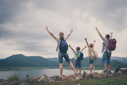 Family with backpacks by a river, hands in the air