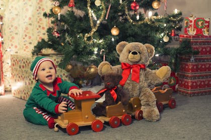 Baby playing with wooden toy train and big teddy bear under Christmas tree