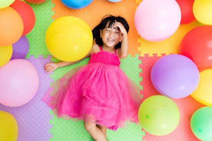 Child in a pink dress laying on the floor surrounded by multicoloured balloons