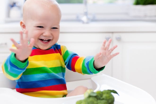 laughing baby sitting in high chair with whole broccoli on the tray 