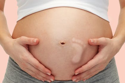 close up of pregnant woman touching her bare tummy with kicking baby footprint over pink background