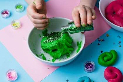 Kid pouring glitter into bowl of green playdough