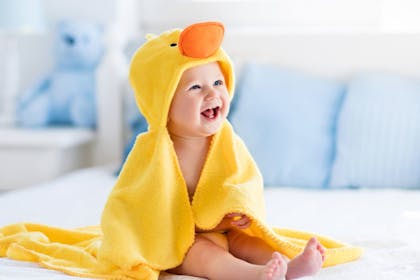 Happy laughing baby wearing yellow hooded duck towel sitting on parents bed after bath or shower. 