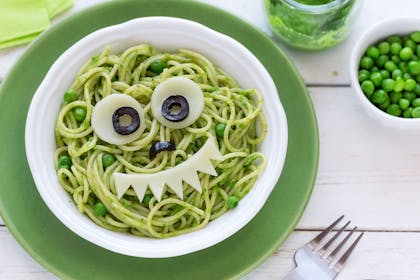 Bowl of pasta with pesto and peas and face made out of cheese and olives