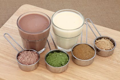 Two protein shakes alongside food powder supplements from left to right: chocolate whey, wheat grass, pomegranate and maca root.