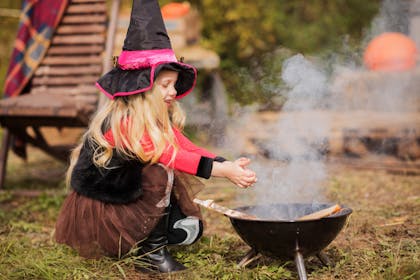 Little girl wears witch hat and costume while pretending to conjure magic over wood burner for Halloween