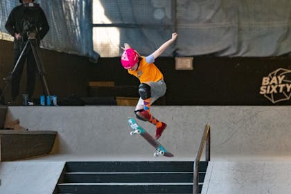 A teenage girl wearing a hot pink helmet performs a huge jump with her skateboard at BaySixty6 skate park 