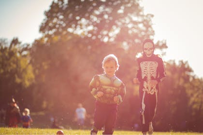 A toddler wearing a Hulk Halloween costume and a taller child wearing a skeleton costume race through a park