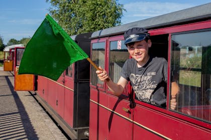 A smiling boy wearing an Engine Driver hat waves a green flag on the Bure Valley Railway, Norfolk