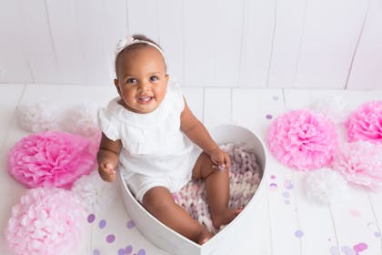 Smiling baby posed sitting in a heart-shaped box and surrounded with pink flowers
