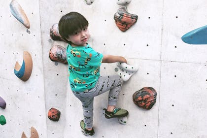 A young child in a t-shirt and joggers turns to smile at the camera while holding on to a climbing wall at The Arch Climbing Wall