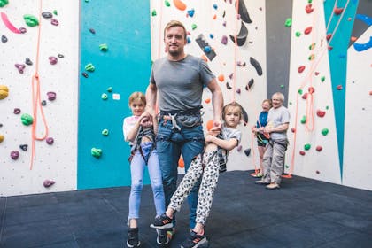 A smiling man carries two young girls by their climbing harnesses at Big Rock Hub, Milton Keynes