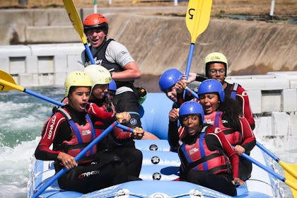A group of teens in protective gear look scared and excited in a blue inflatable boat at the Lee Valley White Water Centre