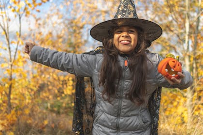 Little girl dressed in duffel coat, witch's cloak and pointy hat poses in woodland with Halloween pumpkin