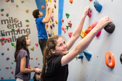 A smiling girl in a black t-shirt holds onto a climbing wall, while two other climbers in the background also enjoy the Mile End Climbing Wall