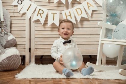 A kid at a party holds a balloon with a birthday banner behind him 