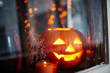 A pumpkin carved into a Halloween jack o'lantern sits lit on a window sill with autumnal ornaments