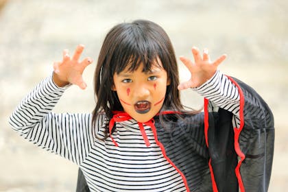 Little girl poses as a vampire for Halloween, dressed in a stripy top, black and red vampire's cloak with lips painted in black face paint 