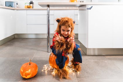 Kid dressed in werewolf costume sat growling on kitchen floor next to Halloween pumpkin lanterns
