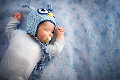 Young baby lying on their back sucking a dummy and wearing a blue knitted owl hat