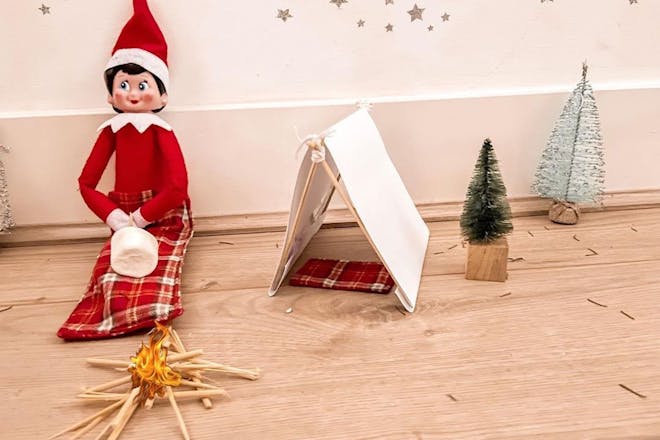 Christmas Elf on the shelf toasts marshmallow on match stick campfire next to model tent