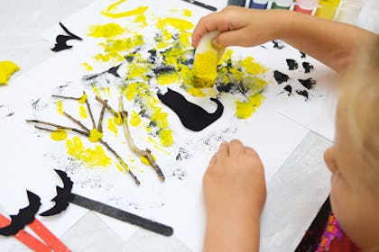A toddler paints a Halloween haunted forest scene using twigs, cat and bat silhouettes cut from black paper, and black and yellow sponge painting