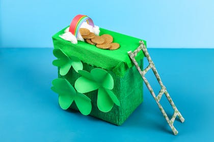 Leprechaun trap made out of cardboard box covered in green paper, paper shamrocks, with a paper rainbow and model ladder