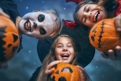 Kids in halloween costumes look down at the camera while holding pumpkins