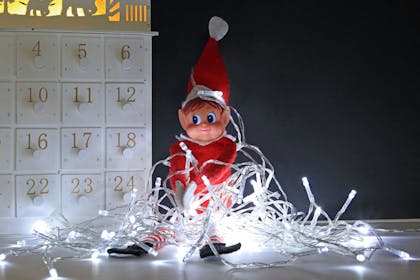 Elf on the Shelf tangled up in Christmas lights next to an advent calendar