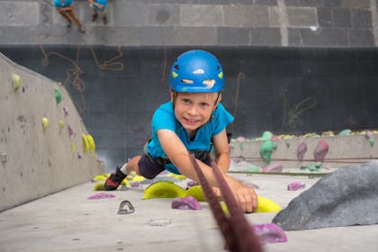 A smiling boy in a blue climbing helmet clings to a wall at Edinburgh International Climbing Arena