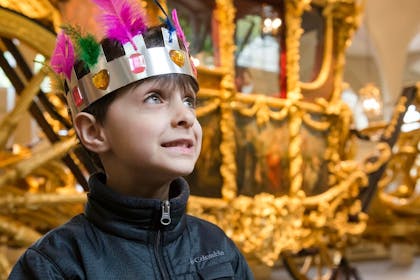 A smiling boy wears a handmade crown with feathers and jewels, standing in front of the Gold State Coach 