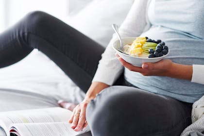 Pregnant woman sitting on bed, eating bowl of fruit and yoghurt and reading book