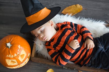 Toddler sleeps on floor next to Halloween pumpkin and wearing witch's hat