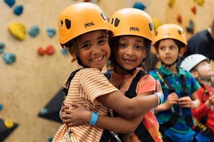 Two young girls wearing climbing helmets hug in front of a climbing wall at Westway Climbing Centre