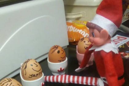 Christmas Elf on the Shelf sits with eggs in egg cups with faces drawn on them