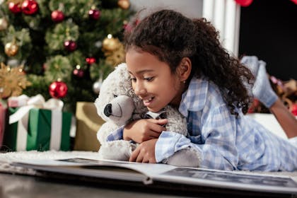 Girl browses photo album in pyjamas while cuddling teddy bear by Christmas tree