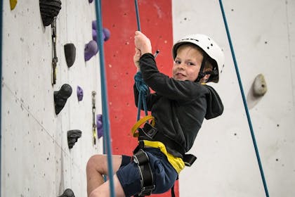 A young boy in a black hoody and a white climbing helmet smiles down from a wall at the Indy Climbing Wall, Anglesey