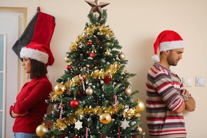 An unhappy couple stand either side of a Christmas tree looking fed up