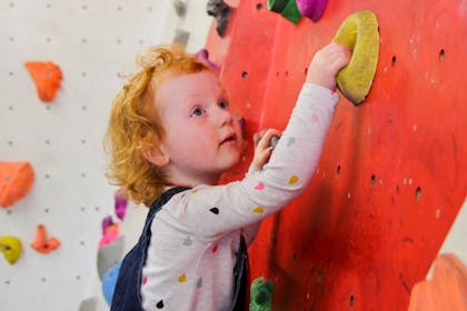 A toddler in dungarees climbs a wall at The Climbing Academy