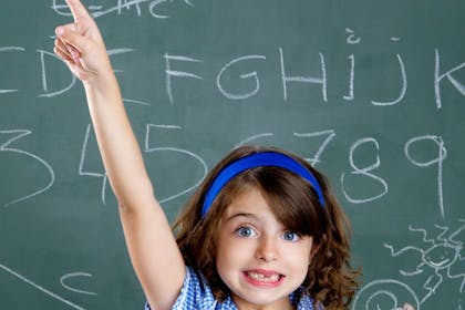 girl in blue with hand up in front of chalk board