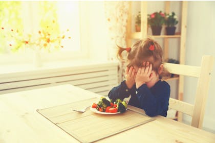 Little girl sitting at a dining table refusing to eat her vegetables