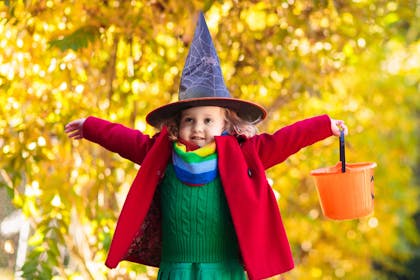 Little girl dressed in witch costume for Halloween stands with arms outstretched while holding orange pumpkin bucket