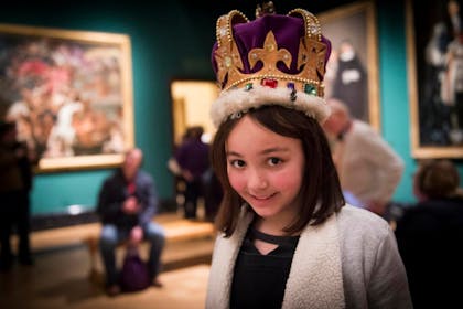 A smiling girl wears a huge purple and gold crown in the Queen's Gallery in London