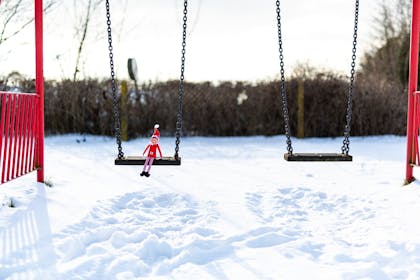 Christmas Elf on the Shelf sits on a swing in a snowy park