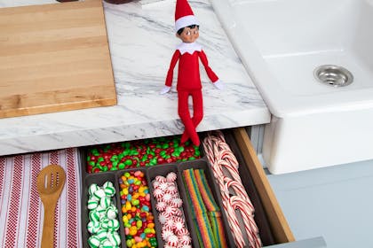 Elf on the shelf sitting near cutlery drawer. Cutlery has been replaced with sweets