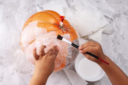 A toddler's hands applying papier-mâché to a pumpkin-shaped balloon for Halloween