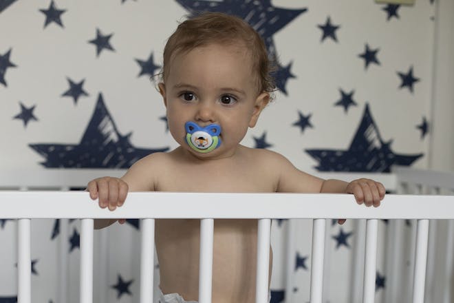 baby with dummy in mouth standing up in white cot